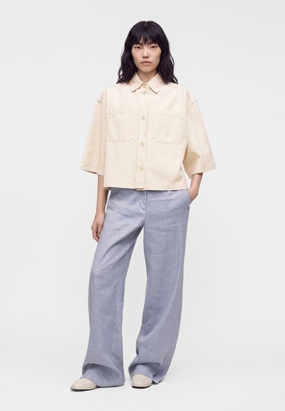 Femme portant une chemise beige ample à boutons avec de larges manches courtes et un pantalon large bleu clair, debout avec une main dans la poche.