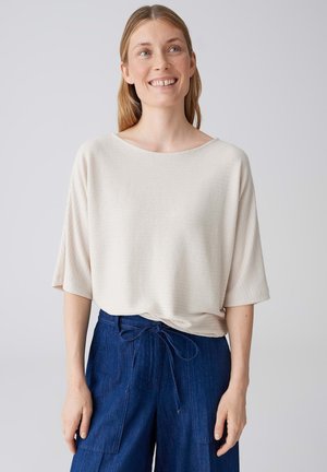 Donna sorridente con capelli castano chiaro che indossa una maglia beige a maniche corte, ampia e dalla texture lavorata, infilata in pantaloni blu navy a vita alta con coulisse frontale.