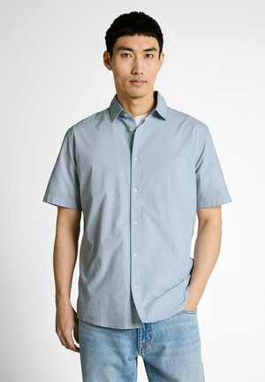 Mann trägt hellblaues, kurzärmeliges Hemd mit Knöpfen und hellblaue Jeans, steht mit einer Hand in der Tasche vor einfarbigem Hintergrund.