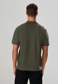 Maslinasto zelena polo majica kratkih rukava s teksturiranim materijalom, klasičnim ovratnikom i otvorenim stražnjim dijelom. Majica ima ravni rub i opušteni kroj.