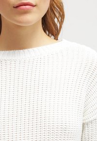 Biały sweter z dzianiny o okrągłym dekolcie, z fakturowanym wzorem i luźnym krojem. Tkanina wydaje się lekka i przewiewna.