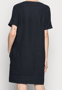 Osoba v volných, krátkých černých šatech, zobrazená zezadu, s malým tetováním čísla 5 na levém ruce.