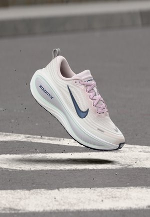 Hele roosa Nike ZoomX tennari tumesinise Swoosh-logoga, hõljuv halli asfaldi kohal valge teepiirde kohal.