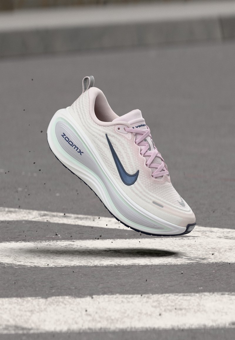 Lyserosa Nike ZoomX-sneaker med marineblå swoosh-logo, svevende i luften over en hvit veikantmarkering på grå asfalt.