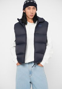 Tumši zilā pufīgā veste ar kapuci, ar izšūtiem posmiem, priekšējiem rāvējslēdzējiem un logotipa detaļām. Apvienota ar baltu kapuci un gaismas ziliem džinsiem.