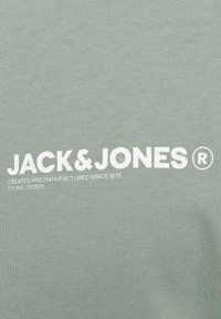 Lichtgroene katoenen T-shirt met witte bedrukte tekst "JACK & JONES" en "GECREËERD EN GEFABRICEERD SINDS 1975," met een soepele textuur.