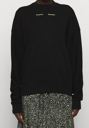 Osoba nosząca czarną bluzę z małym żółtym napisem "Proenza Schouler" oraz spódnicę z czarnymi i beżowymi abstrakcyjnymi wzorami geometrycznymi.