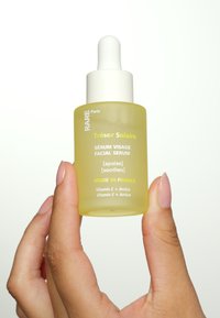 Gul glasflaska med vit pipett, märkt "Trésor Solaire", innehåller ansiktsserum med text på franska. Handen håller flaskan.