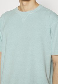 Světle modré bavlněné tričko s kulatým výstřihem. Krátké rukávy, jemná textura a ševové detaily poblíž výstřihu.