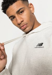 Hoodie gris clair en tissu doux, avec un logo "new balance" brodé en noir sur la poitrine et des cordons assortis.