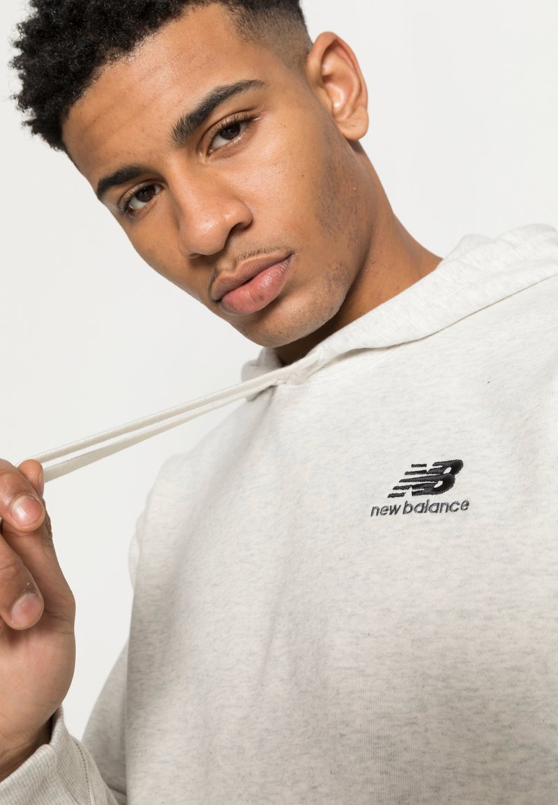 Hoodie grigio chiaro realizzato in un tessuto morbido, con un logo ricamato in nero "new balance" sul petto e cordini coordinati.