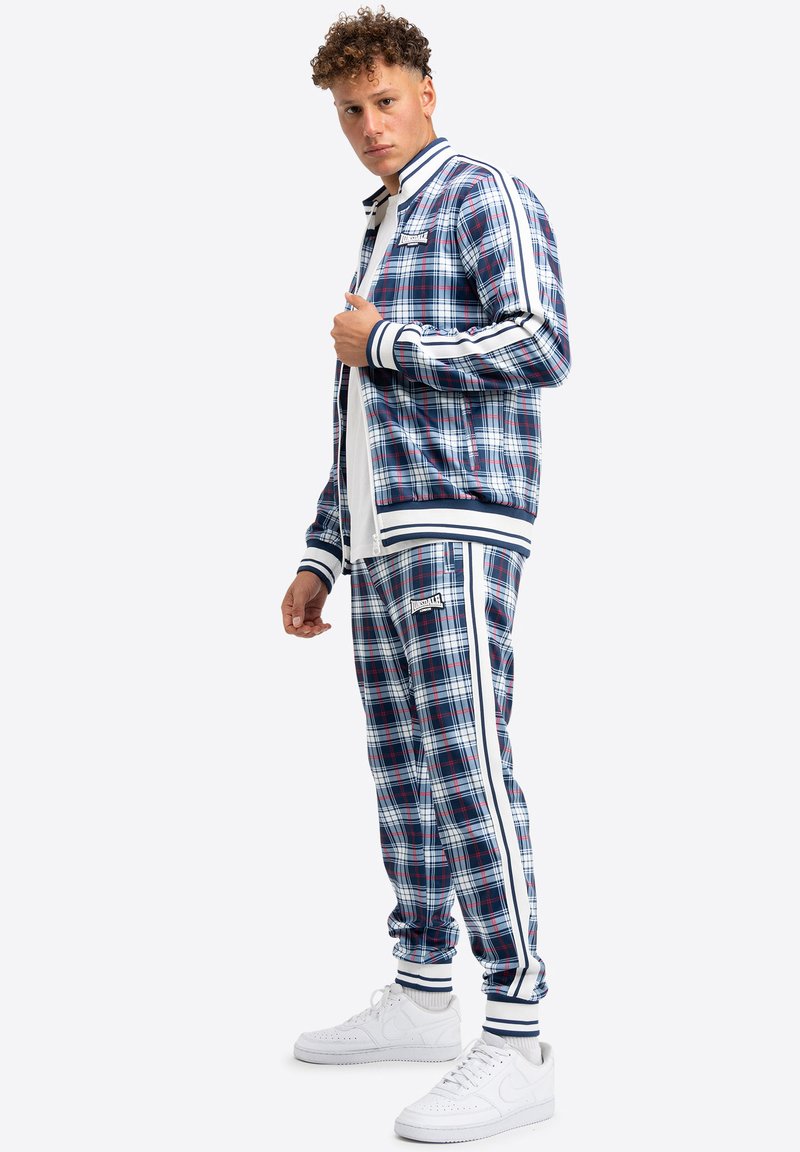 Karierter Trainingsanzug in Marineblau, Rot und Weiß, mit Streifen an den Ärmeln und Beinen, versehen mit gerippten Bündchen, getragen mit weißen Turnschuhen.