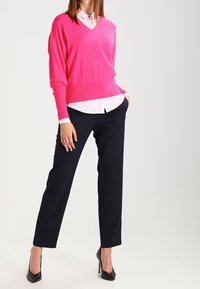 Fuchsia V-Ausschnitt-Pullover über einem weißen Kragenhemd, kombiniert mit schwarz maßgeschneiderten Hosen und schwarzen High Heels.