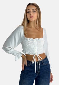 Hvid cropped bluse med smock-detaljer foran, lange puffede ærmer og justerbart bånd i taljen, parret med mørke denim jeans.