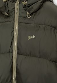 Chaqueta acolchada verde oliva con cierre de cremallera y capucha ajustable. Presenta una textura acolchada y un pequeño logo en el pecho.