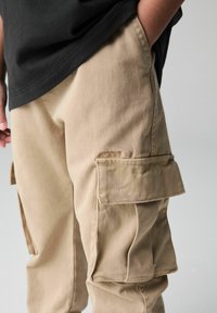Khaki cargobyxor i bomull, med flera fickor, en texturerad yta och subtil branding på låret.