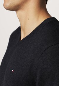 Pull noir à col en V en matière tricotée douce. Présente un petit logo rouge, blanc et bleu sur le côté gauche de la poitrine et des finitions côtelées.