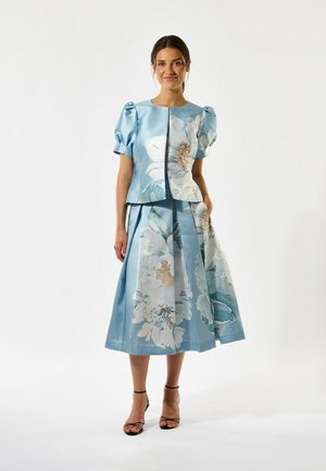 Mujer vestida con una blusa de mangas abullonadas azul claro y una falda midi a juego con grandes estampados florales blancos, de pie y sonriendo, fondo blanco.