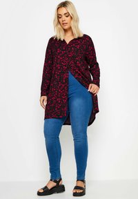 Svart långärmad skjorta med röd blommönstring, knapp-down krage, hög-låg nederkant; matchas med blå tajta jeans och svarta sandaler.