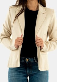 Light beige blazer with a structured design, slim fit, and two side pockets, worn over a black top and paired with blue jeans.