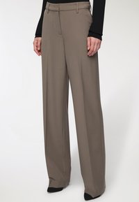 Pantaloni a gamba larga in un tessuto marrone chiaro. Presentano una texture liscia, una parte anteriore sagomata, passanti per la cintura e una chiusura con zip. Indossati con calzature nere.