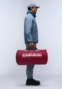 Borsa a tracolla rossa con manici marroni, dotata di un logo e una piccola bandiera. Il modello indossa una giacca blu, pantaloni beige e stivali neri.