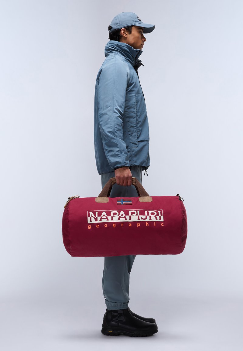 Borsa a tracolla rossa con manici marroni, dotata di un logo e una piccola bandiera. Il modello indossa una giacca blu, pantaloni beige e stivali neri.
