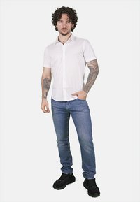 Camicia bianca a maniche corte, slim fit, in cotone. Indossata con jeans denim blu e scarpe da ginnastica nere, con vari tatuaggi visibili.