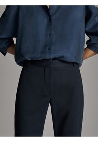 Chemise en satin bleu marine à boutons avec manches retroussées, associée à un pantalon ajusté foncé présentant une texture lisse et des poches avant.