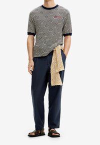 Námořnická modrá a krémová krátký rukáv pletené tričko s geometrickým vzorem, v kombinaci s tmavými kalhotami. Béžová pletená šála uvázaná v pase; nošeno s černými sandály.