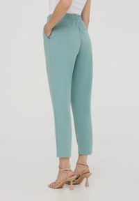 Pantaloni a sigaretta verde menta con cintura elasticizzata, tasche laterali e texture liscia, abbinati a sandali con tacco beige chiaro.