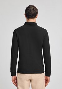Zwarte polo met lange mouwen en een klassieke kraag, gemaakt van gestructureerde stof. Heeft aansluitende mouwen en een rechte zoom op de taille.