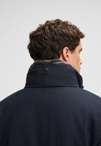 Cappotto blu navy con colletto alto foderato in morbido grigio. Un bottone impreziosisce il colletto, conferendo un aspetto strutturato.