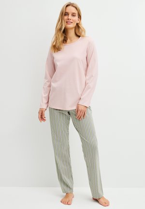 Donna in piedi scalza che indossa una maglia a maniche lunghe rosa chiaro e pantaloni a righe verticali verdi e bianchi su uno sfondo bianco uniforme.