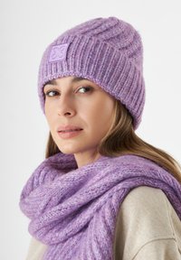 Paarse gebreide beanie met ribbeltextuur en een vierkante logopatch, gecombineerd met een bijpassende chunky sjaal in een vergelijkbare tint.