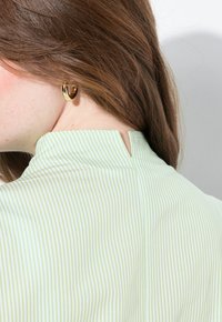 Tessuto a righe verde e bianco con colletto alto, caratterizzato da una fessura sul retro e una texture liscia. Orecchino circolare dorato visibile in primo piano.