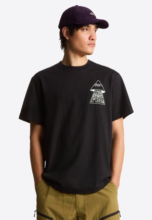 Młody mężczyzna w czarnej koszulce z nadrukiem i fioletowej czapce, z rękami w kieszeniach oliwkowych spodni, patrzący w prawo na jednolitym białym tle.