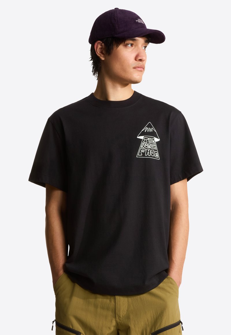 Mladý muž v černém tričku s potiskem a fialové čepici, s rukama v kapsách olivových kalhot, hledící doprava na jednobarevném bílém pozadí.