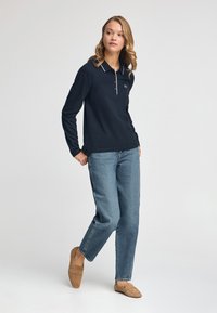 Granatowa koszulka polo z długim rękawem, białym kołnierzykiem i zapięciem na trzy guziki, w połączeniu z jasnoniebieskimi jeansami prostymi i beżowymi butami typu slip-on.