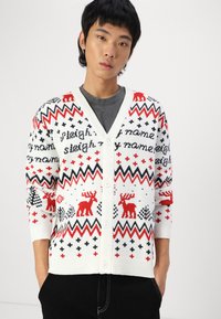 Hvid cardigan med røde rensdyr og sorte accenter, prydet med zigzag-mønster og tekst. Knapper foran og ribstrikkede manchetter.