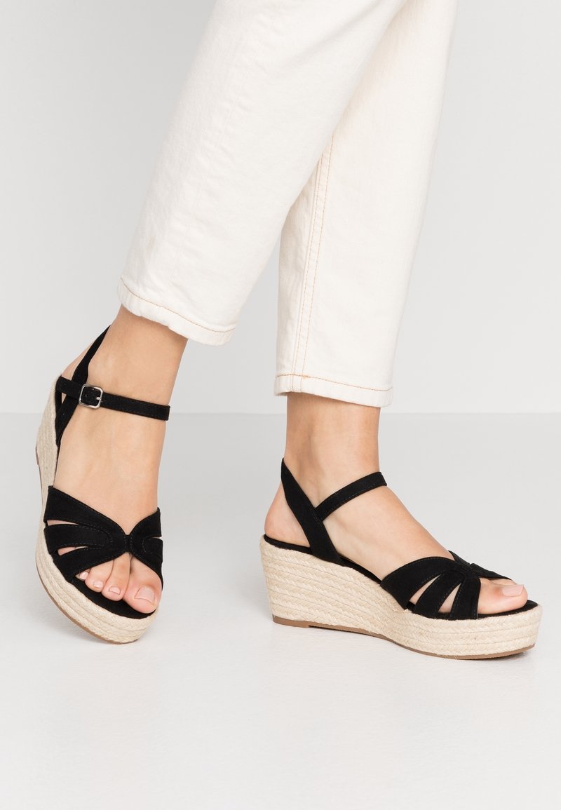Pieds portant des sandales compensées en suède noir avec des brides à la cheville et des talons plateformes tissés clairs, associées à un pantalon blanc raccourci.