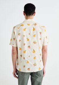 Camisa de manga corta en tejido beige claro con un patrón de ilustraciones de naranjas y fresas. Corte cómodo con cuello y textura suave.