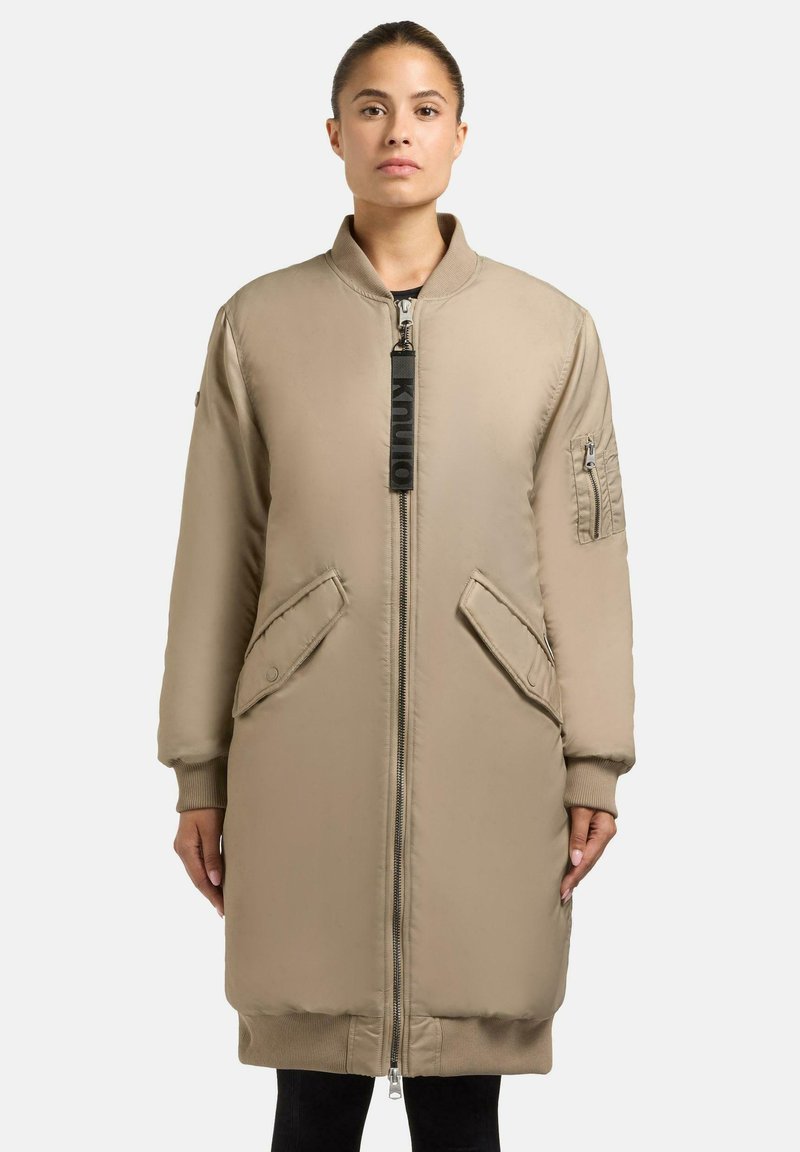 Beige gewatteerde jas met een hoge kraag, ritssluiting aan de voorkant en twee grote zijzakken. Voorzien van ribgebreide manchetten en een gewatteerde patchzak op de mouw.