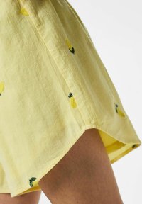 Short en tissu jaune avec de petits citrons brodés porté sur la cuisse d'une personne sur fond blanc.