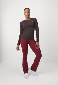 Ilgi piedurkņu brūns tops un bordo cieši pieguļošas bikses. Apģērbs ir ar gludu tekstūru un kontrastējošām krāsām, komplektā ar baltām sporta kedām.