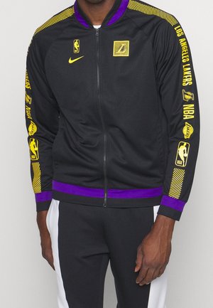 Czarna kurtka na zamek Los Angeles Lakers z żółtymi logo NBA i napisem na rękawach, fioletowy kołnierz i dół, noszona przez osobę.