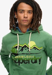 Groene hoodie met bergafbeelding, zachte stof, verstelbare koordjes en voorkantzak. "Superdry" logo in vet zwart lettertype.