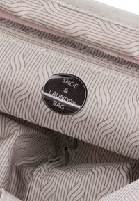 Borsa per scarpe e biancheria in un tessuto beige chiaro con sottili motivi ondulati, un'etichetta circolare nera e un accento di cerniera rosa.