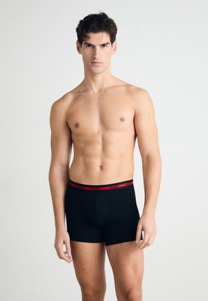Schwarze Boxerbriefs mit einem roten Logo-Bund, die einen eng anliegenden Schnitt und eine glatte Textur aufweisen, präsentiert an einem Modell vor einem neutralen Hintergrund.
