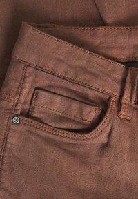 Tela de mezclilla marrón con una superficie texturada, que presenta una cinturilla visible, un pequeño bolsillo y un detalle de remache metálico.